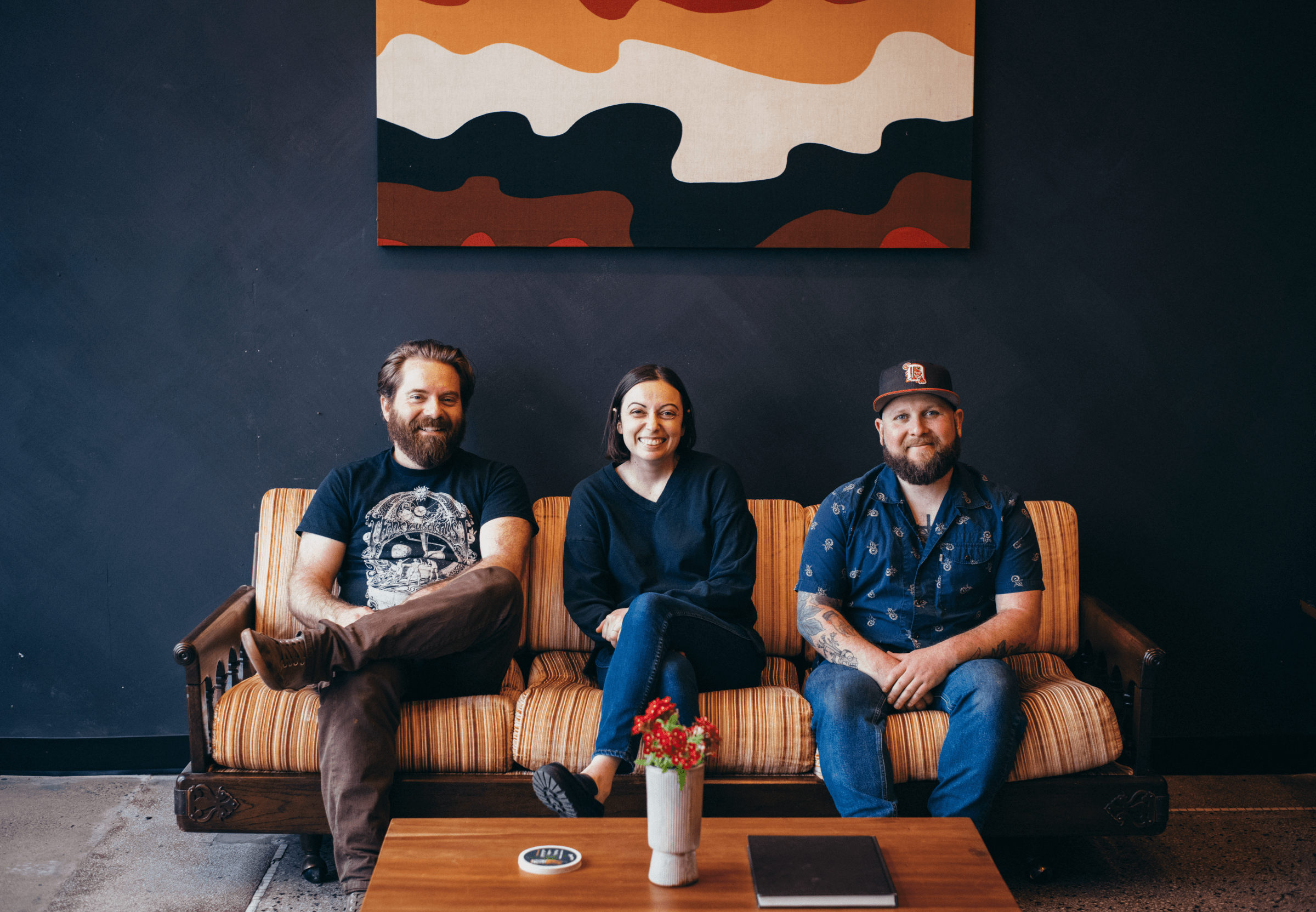Three people sit on a 1970s-style couch against a dark blue wall. 